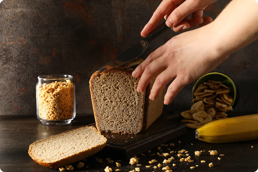 Hands slicing a rustic loaf to measure how many carbs are in a slice of bread.